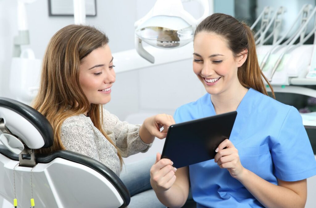 A female dentist in blue scrubs and a woman sitting in a dentist's chair looking at a tablet.