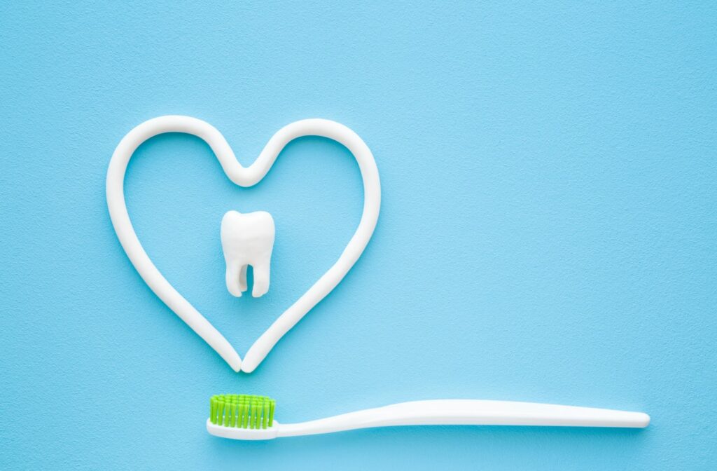 A toothbrush with green bristles beneath some toothpaste that is laid out in the shape of a heart. There is also a plastic model of a tooth in the middle of the heart shape.