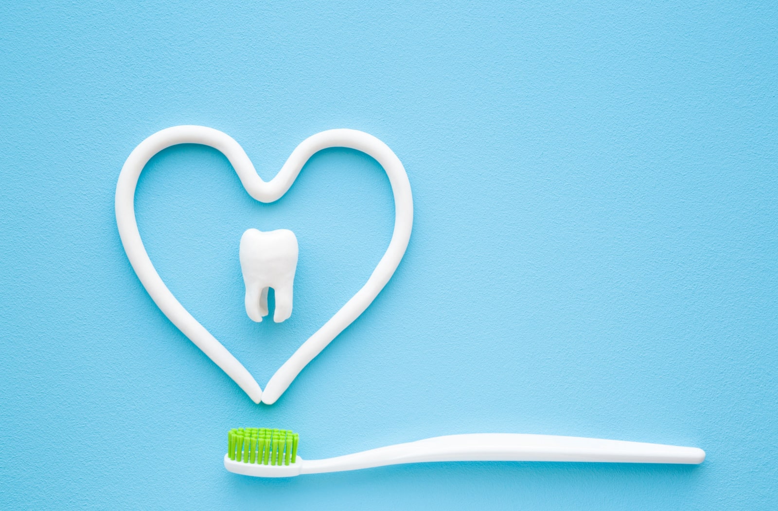 A toothbrush with green bristles beneath some toothpaste that is laid out in the shape of a heart. There is also a plastic model of a tooth in the middle of the heart shape.