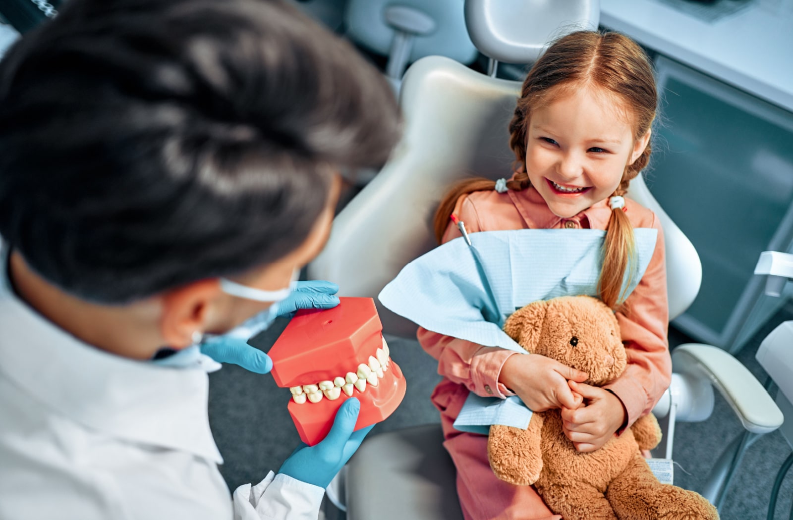 A dentist showing a model of a teeth to a happy child holding a stuffed animal.
