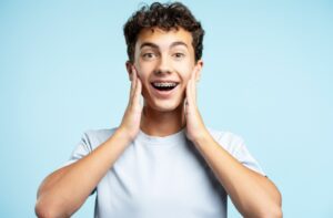 A young man with braces smiles and touches his cheeks.