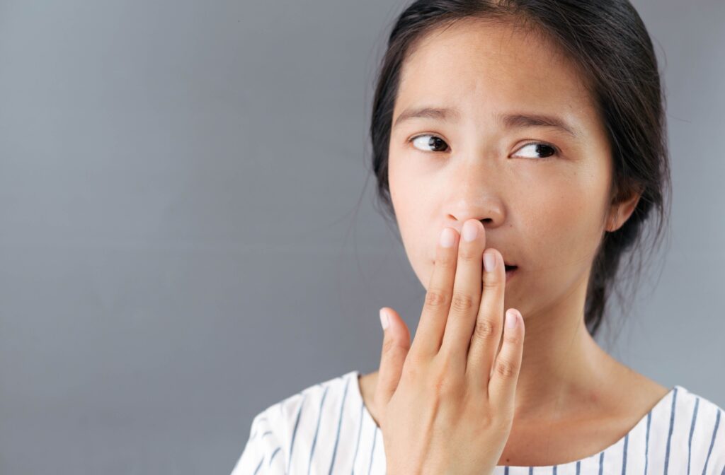 Concerned woman covering her mouth with her hand, due to bad breath, standing against a neutral grey background.