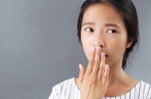 Concerned woman covering her mouth with her hand, due to bad breath, standing against a neutral grey background.