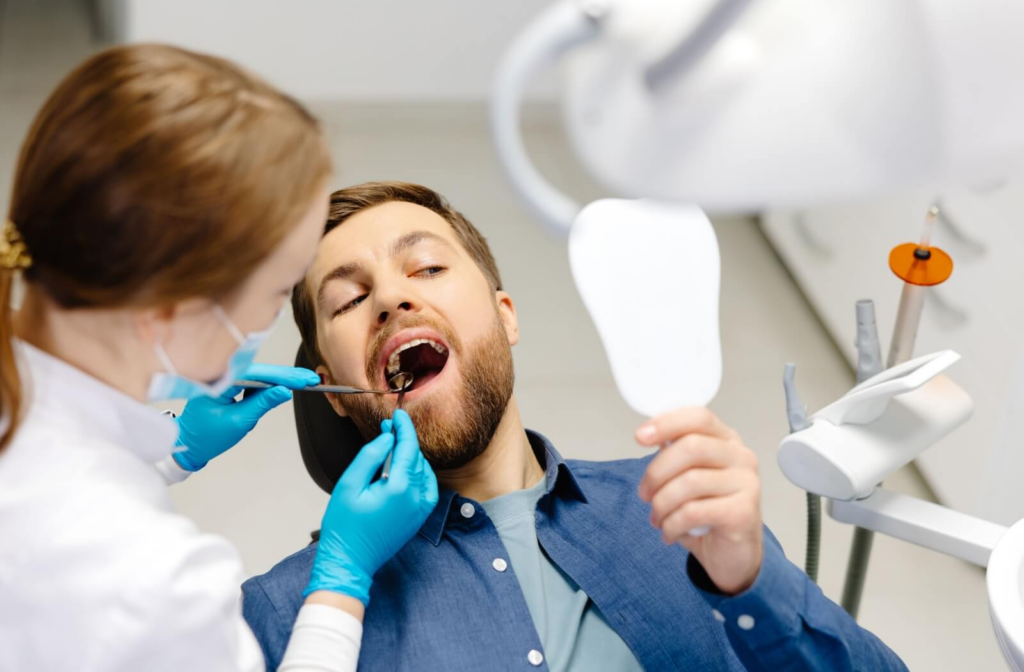 Dentist examining a male patient’s teeth while he holds a small mirror during a dental appointment